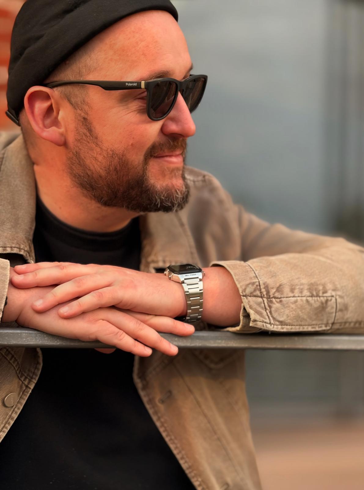 Man wearing a beanie and sunglasses, leaning on a railing outdoors.